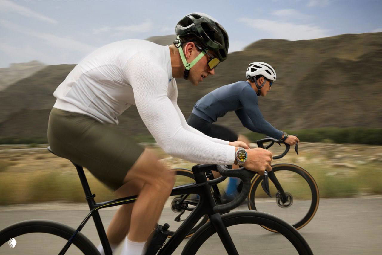 Два велосипедиста на дороге, один в переднем плане с видимыми умными часами на запястье, съемка в движении на фоне холмов.