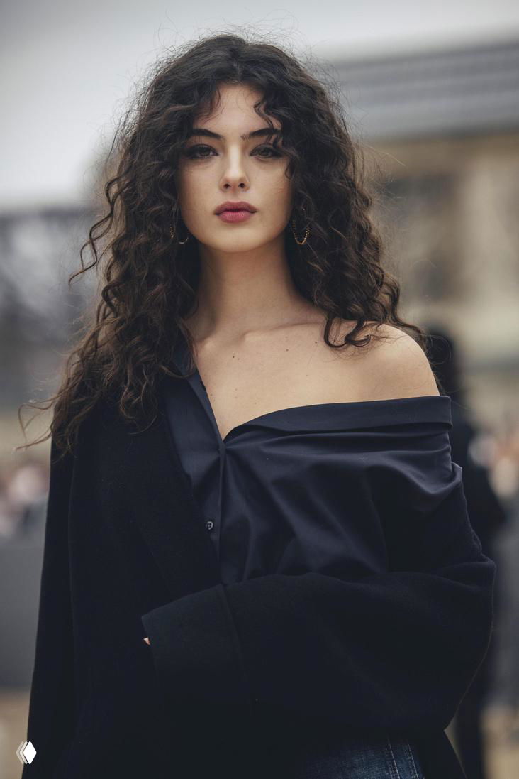 Крупный портрет молодой женщины с волнистыми тёмными волосами на городской улице, расслабленный взгляд и естественный макияж.