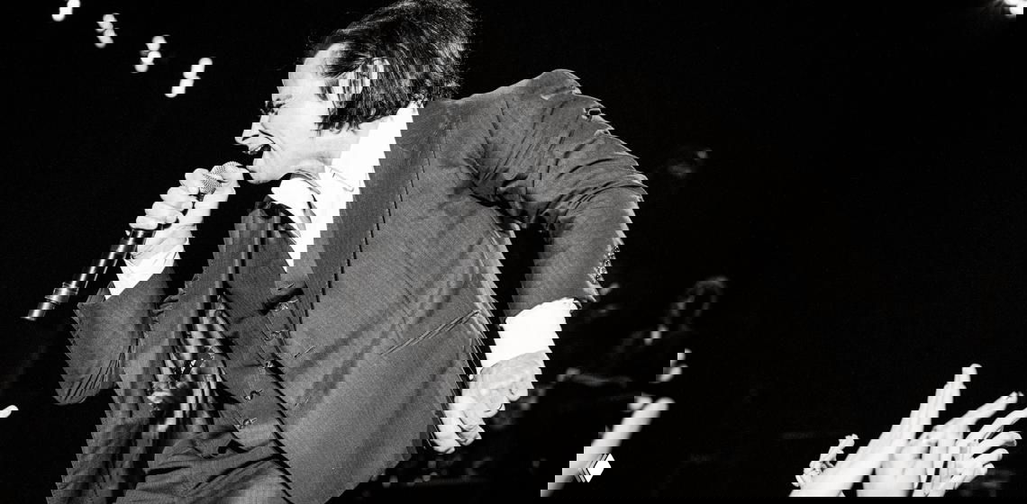 Панорамный чёрно-белый снимок со сцены: исполнитель в костюме наклоняется к зрителям и держит микрофон, видны руки в фан-зоне