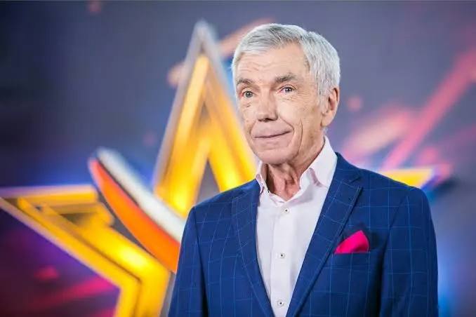 Юрий Николаев в телевизионной студии: пожилой ведущий в синем пиджаке и белой рубашке на фоне яркой звезды и декораций