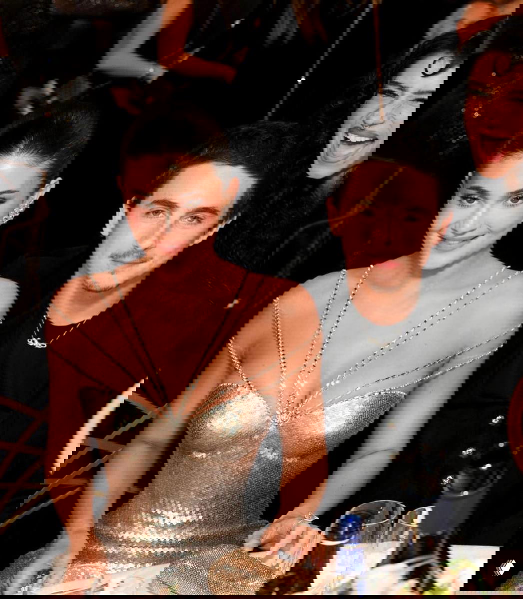 Праздничный кадр за столом: женщина в блестящем золотистом платье сидит рядом с мужчиной, вокруг праздничная сервировка и бутылки вина.