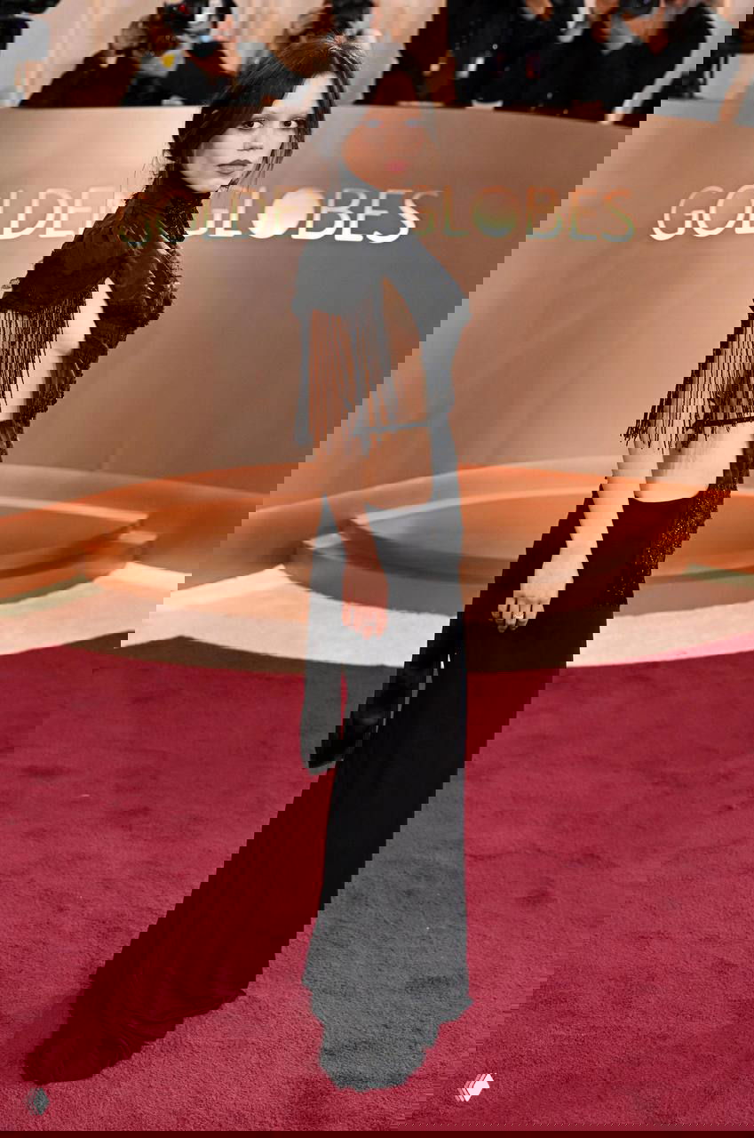 Женщина в чёрном платье с бахромой и заметными вырезами позирует у брендированного фона «Golden Globes», кадр с красной дорожки.