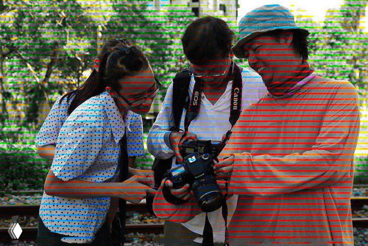 Трое человек на улице внимательно смотрят на экран зеркальной камеры; в кадре показана оптическая иллюзия — цветные полоски на чёрно‑белом фоне.