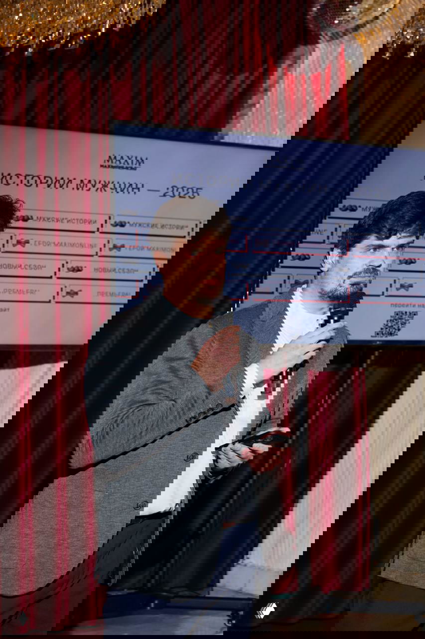 Мужчина в сером пиджаке держит микрофон на сцене вечера запуска проекта, на заднем плане экран с логотипом Maxim и красные шторы