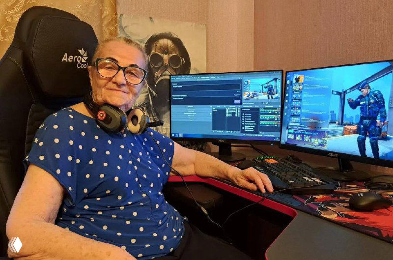 Семидесятилетняя женщина в игровой комнате за многомониторной установкой с клавиатурой и гарнитурой, позирует за компьютером во время стрима.