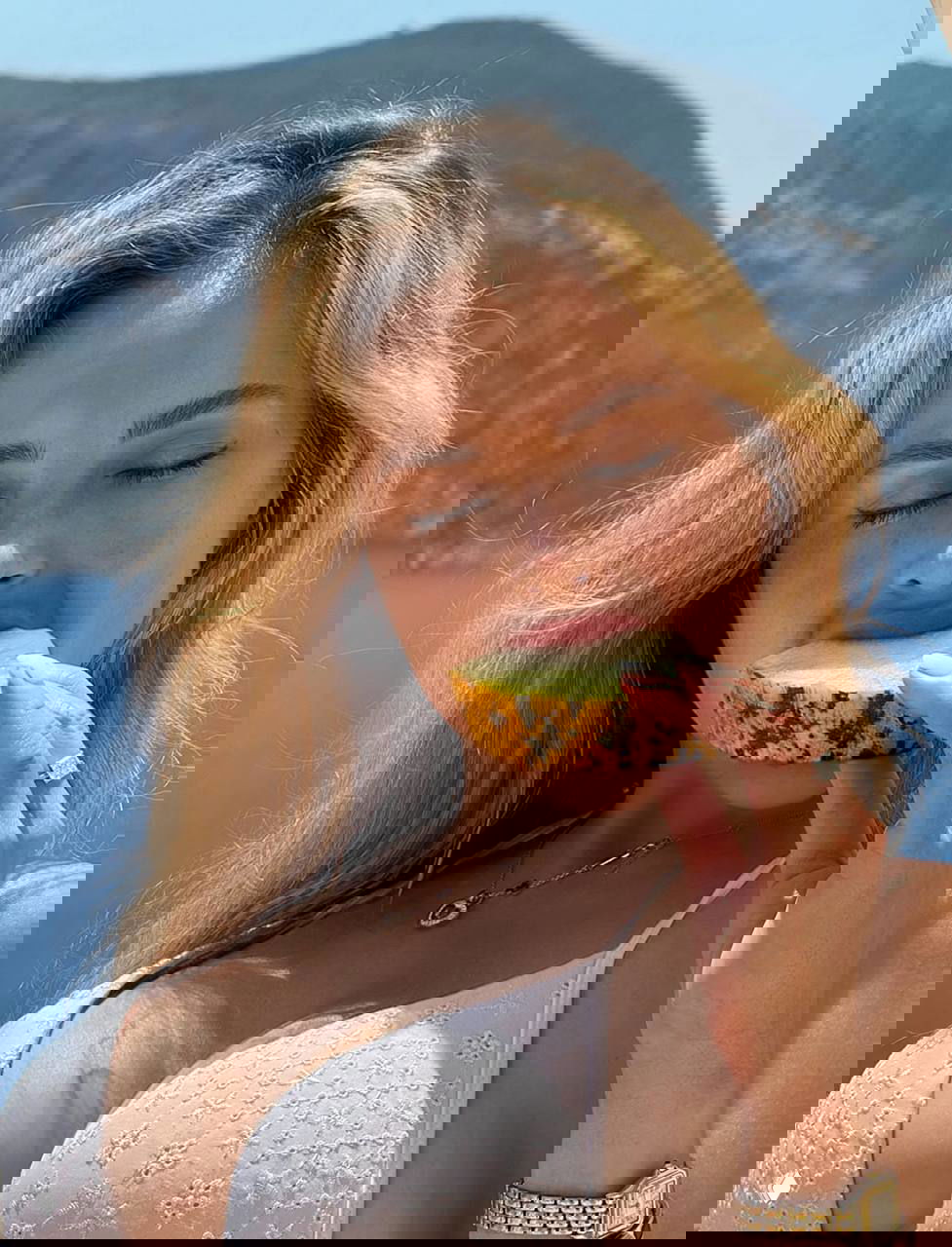 Крупный план женщины в купальнике, она ест дольку дыни у моря; видна верхняя часть торса, лицо с закрытыми глазами и горный фон.