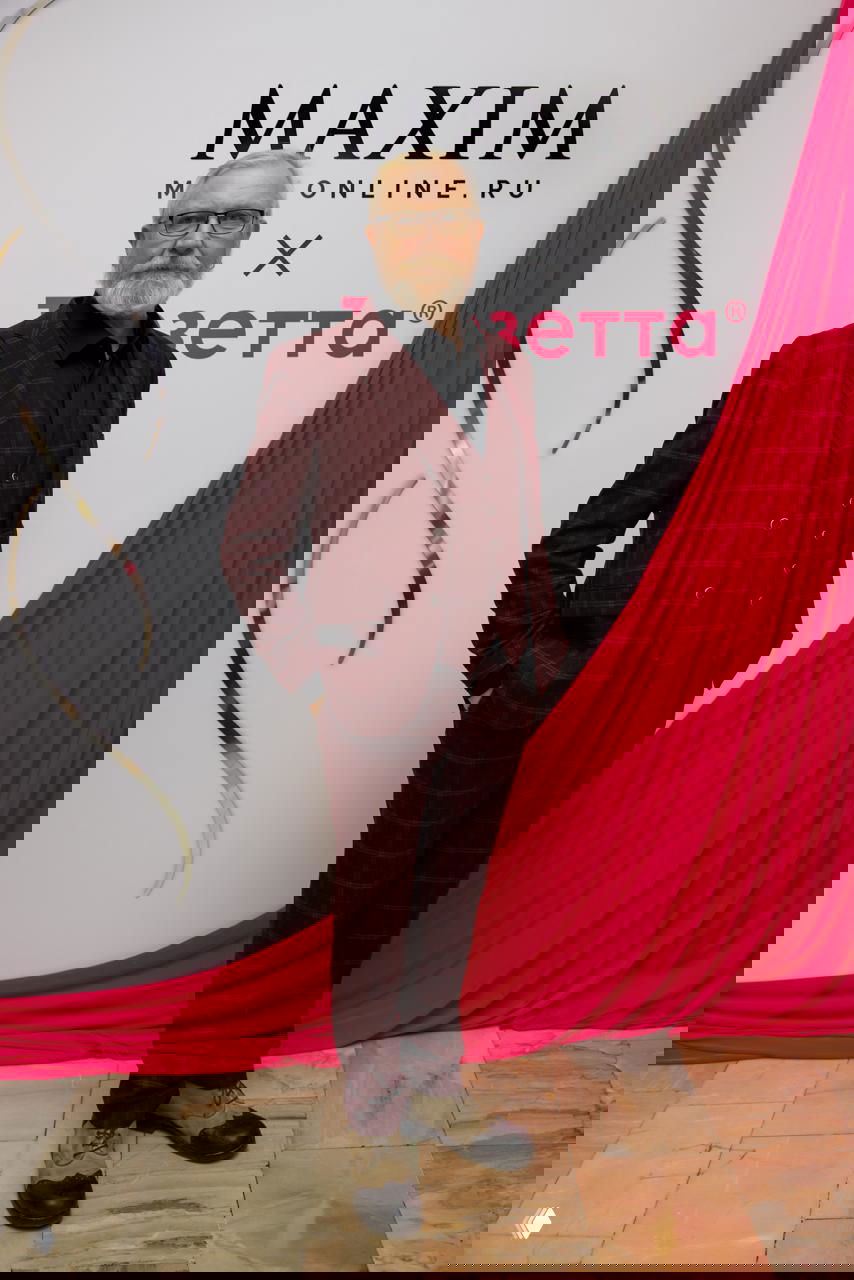 Взрослый мужчина в бордовом костюме позирует на фотозоне с логотипом Maxim, атмосферный кадр красно-розовой декорации и пола