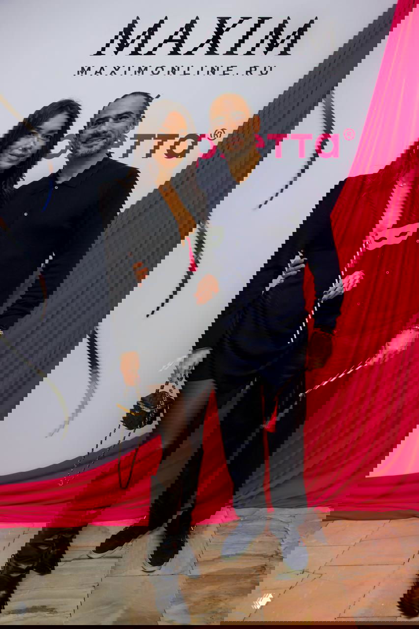 Двое гостей мероприятия позируют на фоне баннера Maxim: женщина в черном платье и мужчина в тёмном свитере, держат маску-аксессуар