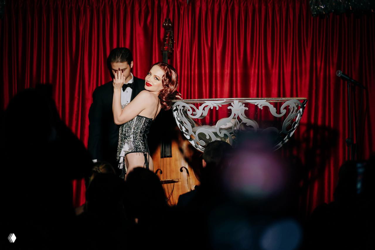 Артистка в блестящем костюме выступает на сцене с красными шторами, в кадре видна часть музыкальной сцены и зрительный зал в затемнении