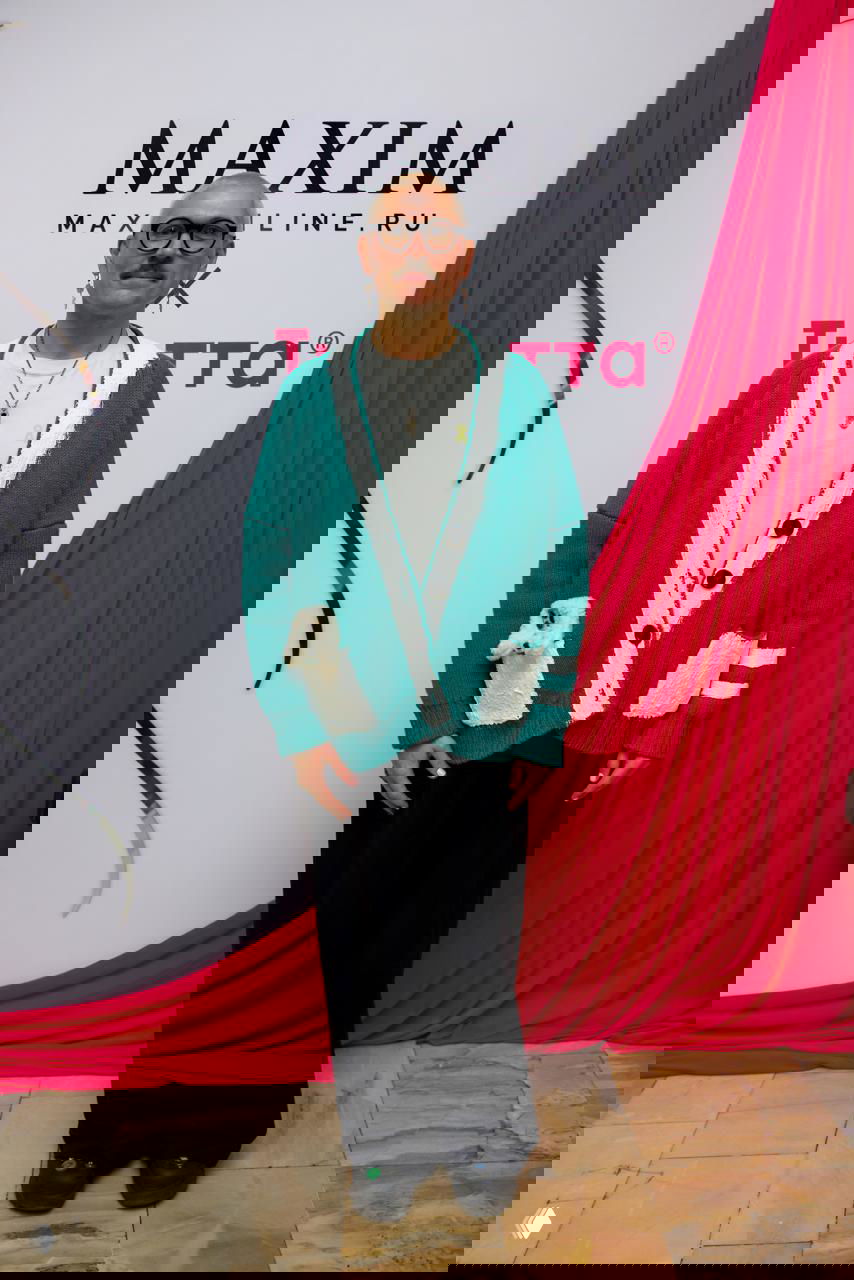 Мужчина в зелёном кардигане позирует у брендированной фотозоны Maxim на фоне яркой розовой драпировки и логотипа мероприятия