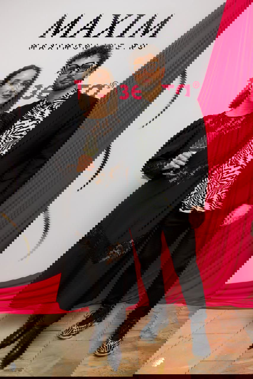 Пара гостей позирует в темной одежде на фоне баннера Maxim, женщина держит фужер с вином, атмосфера коктейльного приема и фотозоны