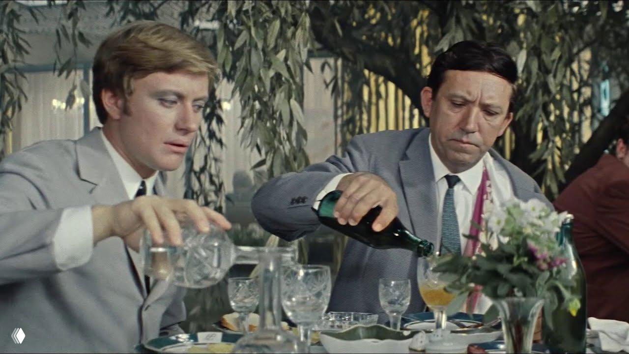 Кадр застолья: двое мужчин в костюмах на фоне сервированного стола наливают алкоголь в бокалы — сцена из фильма/праздника.