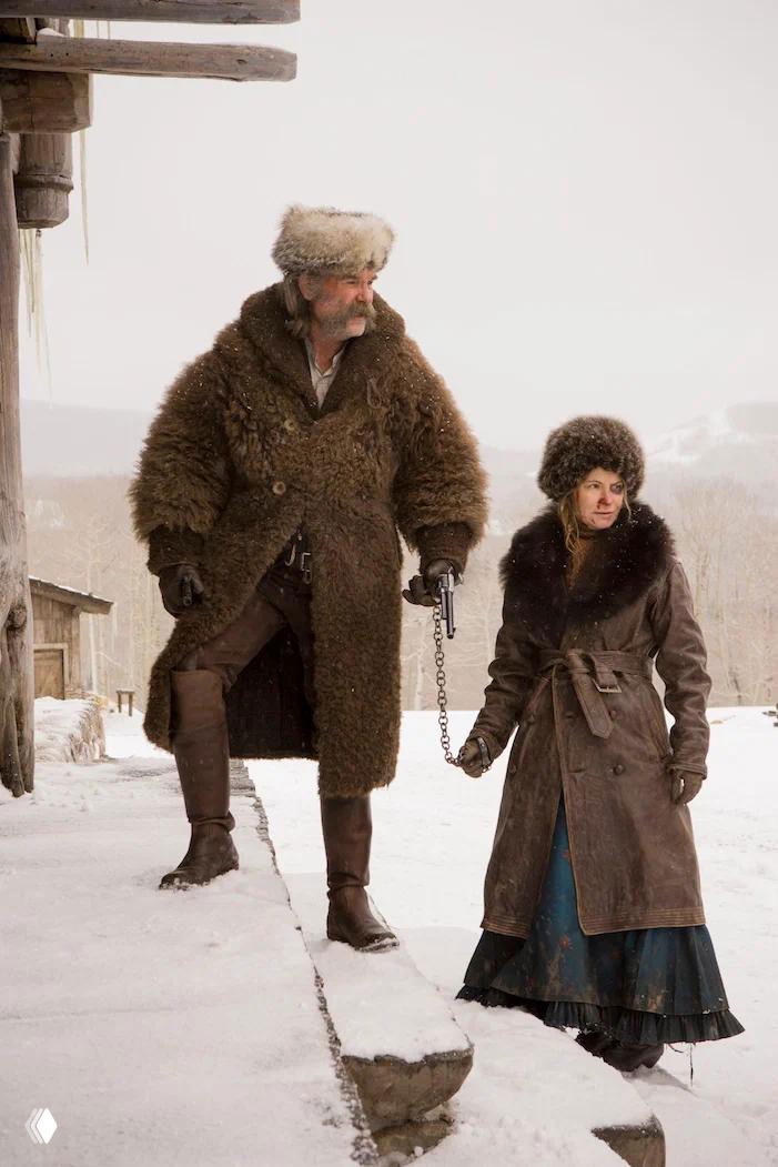 Два персонажа в зимней обстановке у деревянного дома: мужчина в длинной шубе с оружием и женщина в меховом пальто на заснеженной дороге.