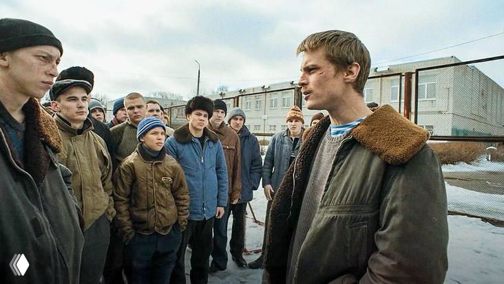 Кадр из сериала: молодой герой стоит лицом к группе подростков на фоне индустриальной застройки и снега, зимняя уличная сцена.