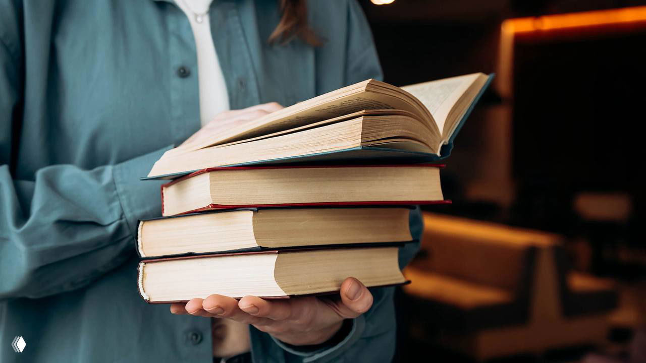 Человек, держащий стопку печатных книг в руках в интерьере с тёплым освещением; крупный план, мягкий фон и логотип в углу.