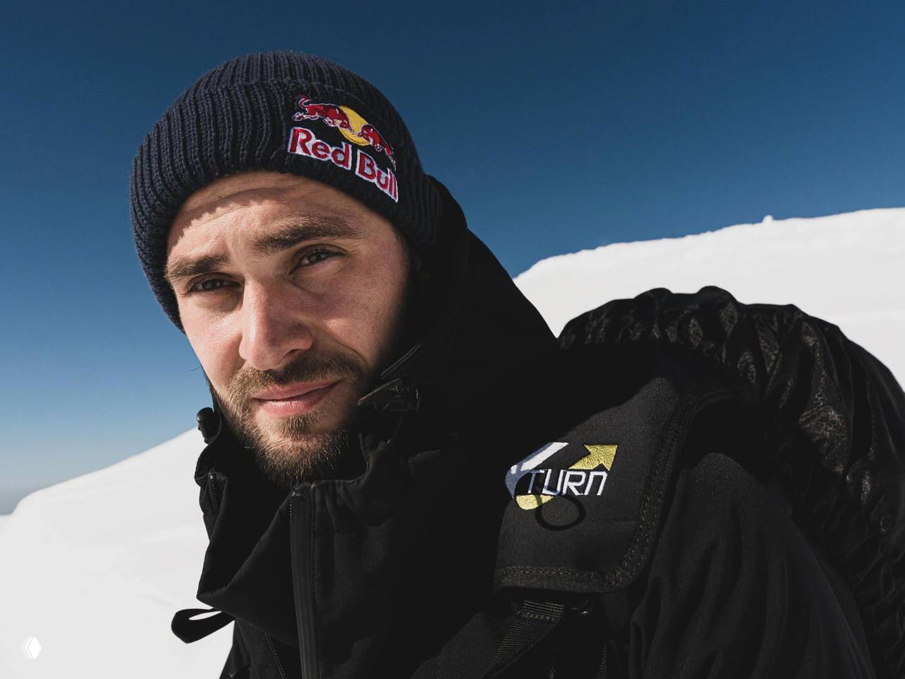 Крупный портрет мужчины в шапке Red Bull на снежной вершине: Егор Посохин в тёплой экипировке, видно лицо, щетину и часть снаряжения.