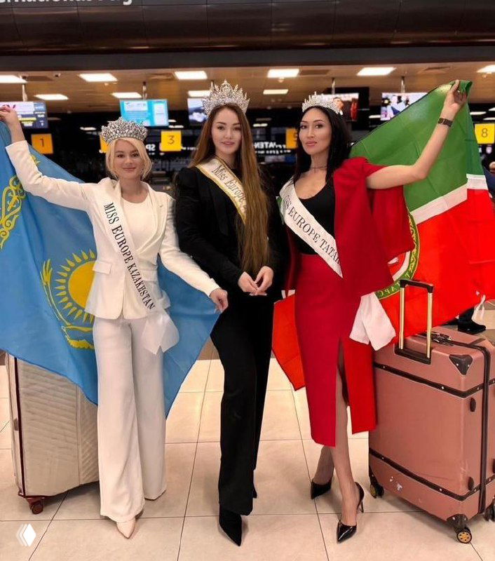 Три конкурсантки в аэропорту с коронами и лентами, позируют с флагами Казахстана и Татарстана рядом с чемоданами; сцена отправления и поддержка.