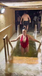 Женщина в красном купальнике поднимается из проруби по ступенькам внутри помещения; вокруг люди и тёплое освещение, кадр из видео.