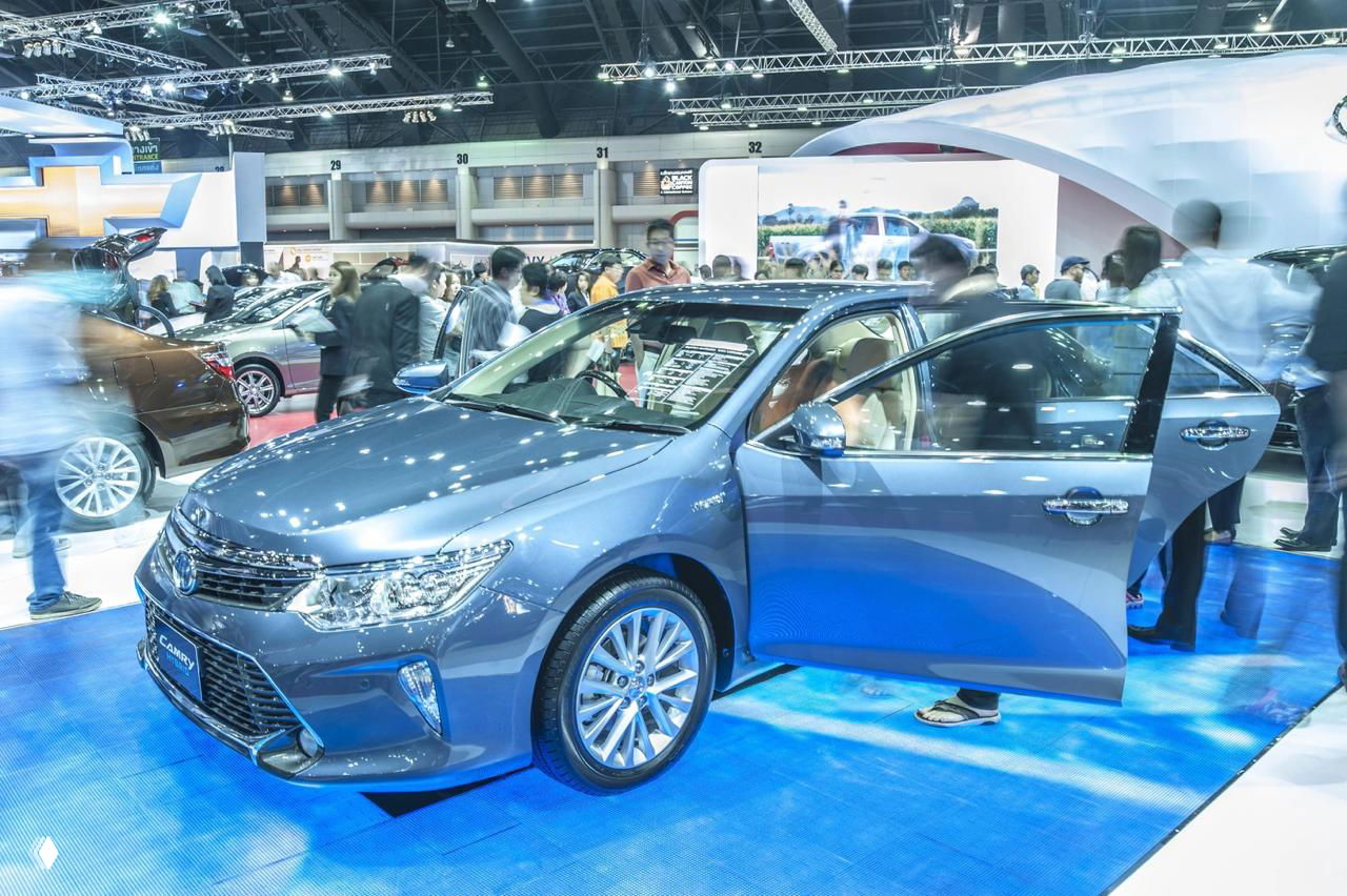 Фото салонного стенда с седаном на синем подиуме в выставочном зале: машина с открытой дверью и посетители вокруг, сцена автосалона.