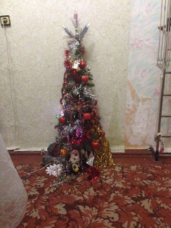 Невысокая ёлка с плотной мишурой и гирляндами, у её основания мягкая игрушка и простые украшения — домашний праздничный уголок.