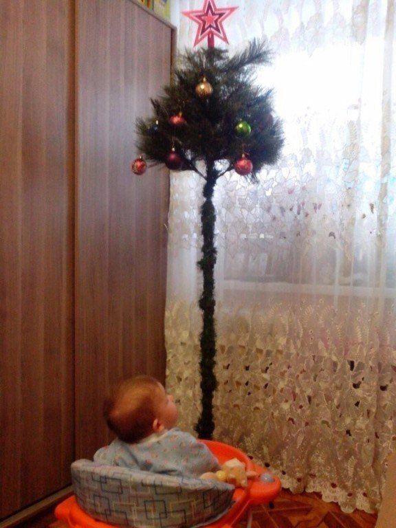 Тонкая высокая ёлка на стойке у окна; рядом ребёнок в ходунках наблюдает за украшениями — бытовой семейный снимок новогодней ночи.