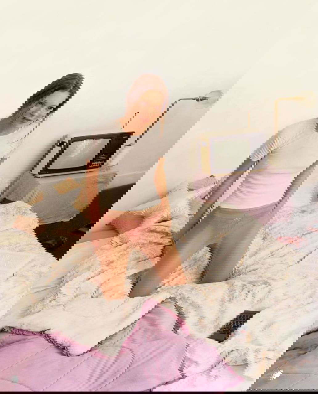 Молодая модель в белой футболке и прозрачных розовых колготках сидит на неубранной кровати рядом с ретро-телевизором и покрывалом.