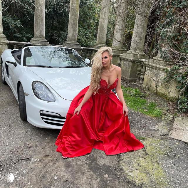 Женщина в длинном красном вечернем платье сидит рядом с белым кабриолетом у каменной ограды и колонн, поза для гламурной фотосессии на открытом воздухе.