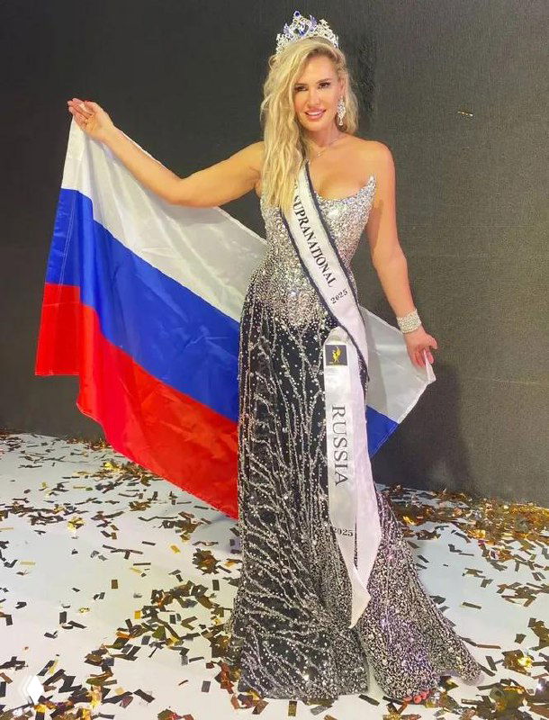 Женщина в блестящем вечернем платье и короне держит российский флаг и ленту с надписью «Russia», стоит на сцене в окружении конфетти после церемонии награждения.