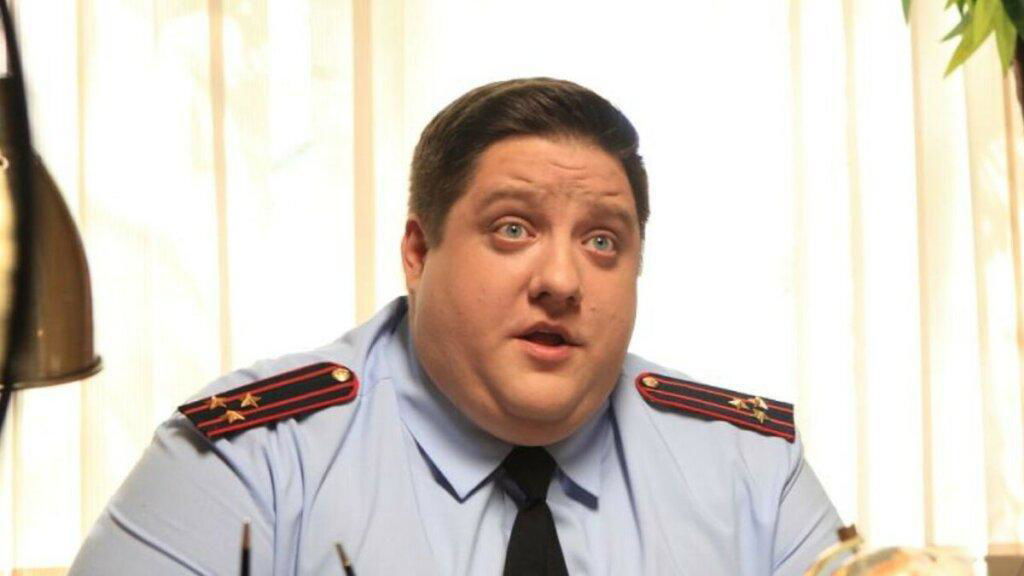 Роман Попов в образе полицейского — крупный план актера в форме за столом, выразительное лицо и светлый фон окна за спиной, кадр со съёмочной площадки.