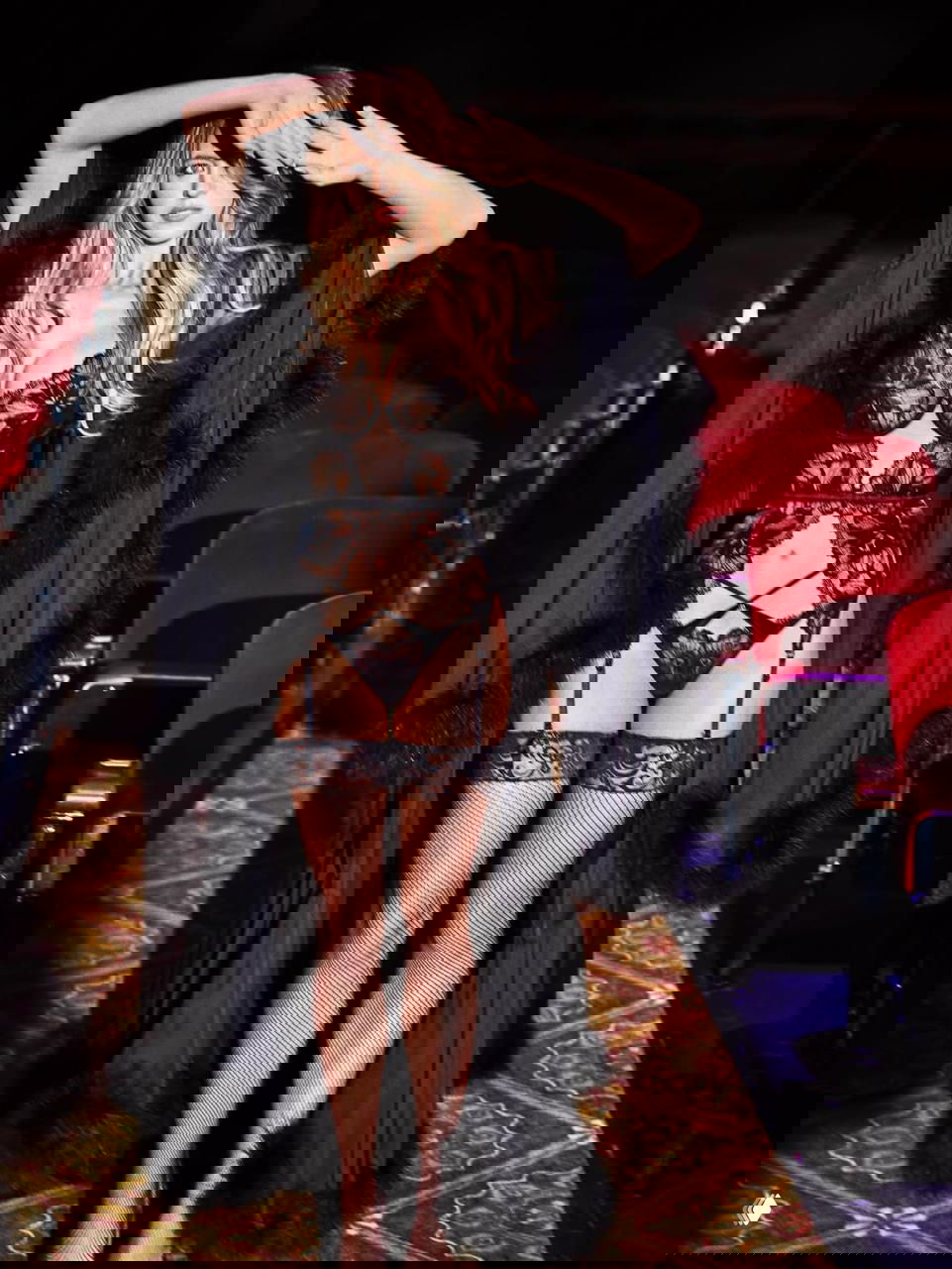 Фотография полного роста Сидни Суини в кружевном наряде, чулках и плаще на театральной сцене; видны красные кресла и гламурная постановка для рекламной сессии.