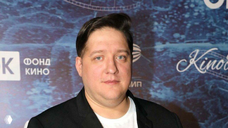 Портрет Романа Попова на кинофестивальной площадке — средний план в деловом пиджаке, нейтральный фон с логотипами и кинофонда.