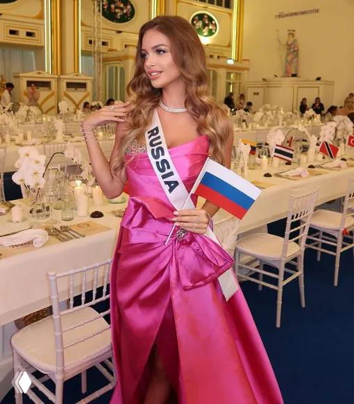 Участница от России в розовом платье с лентой «RUSSIA» и небольшим флажком позирует в банкетном зале на фоне сервированных столов.