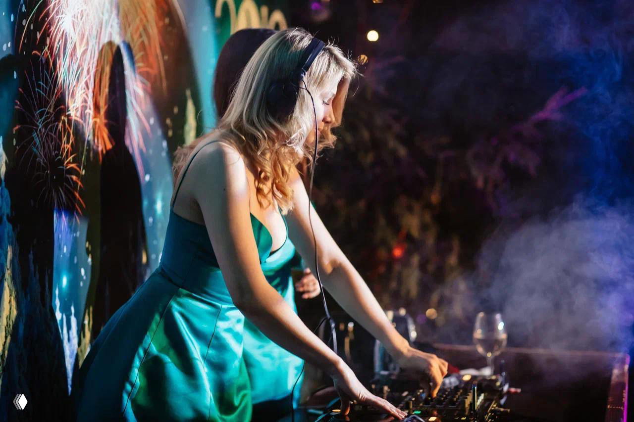 Ди-джей в бирюзовом платье играет за пультом на вечернем шоу; огни, дым и праздничная музыкальная атмосфера.