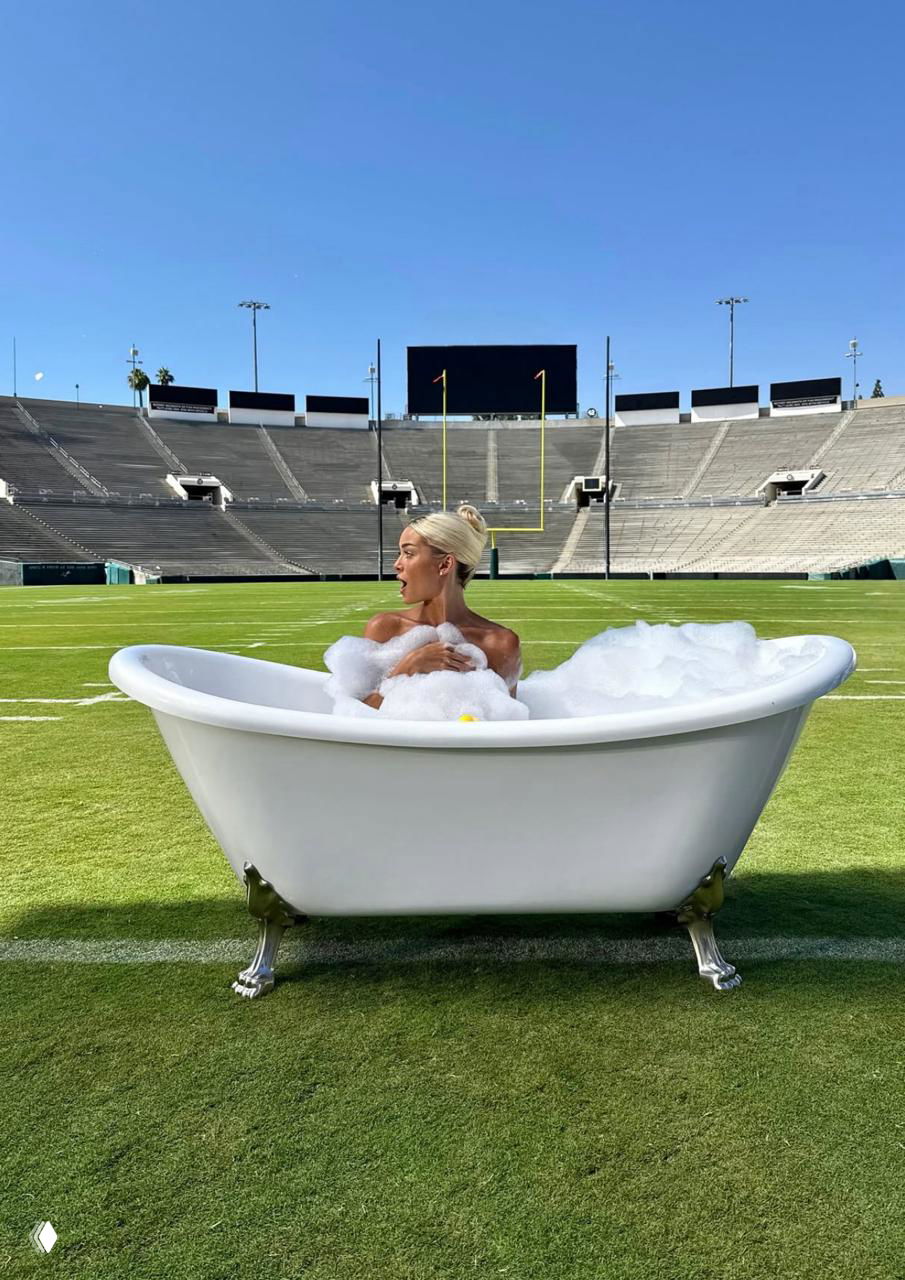 Женщина в ретро-ванне с пеной, установленной посреди зелёного футбольного поля; пустые трибуны, табло и ворота видны на заднем плане.