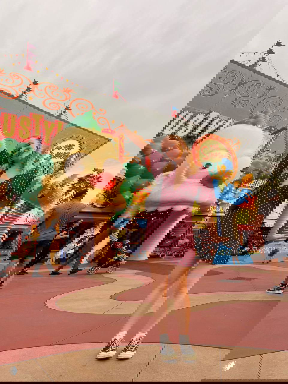 Женщина в летнем платье позирует возле крупной мультяшной фигуры и аттракционов в парке развлечений; вокруг посетители парка и красочные декорации.