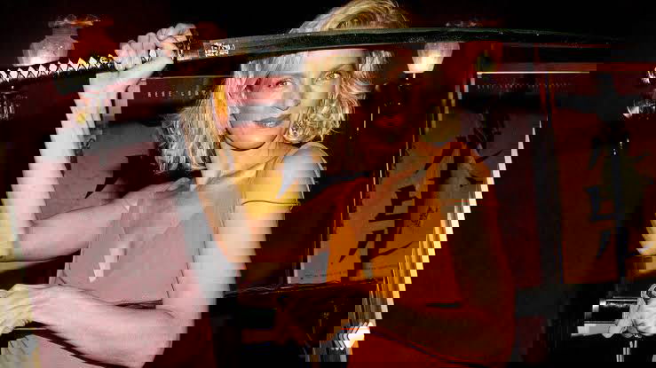 Женщина в оранжевом платье держит длинный предмет над плечом на сцене; фото сделано в вечерней обстановке, видно часть декора.