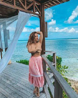 Анфиса Чехова на деревянной веранде у моря в длинной розовой юбке, прикрывает грудь рукой на фоне бирюзовой воды и облачного неба.