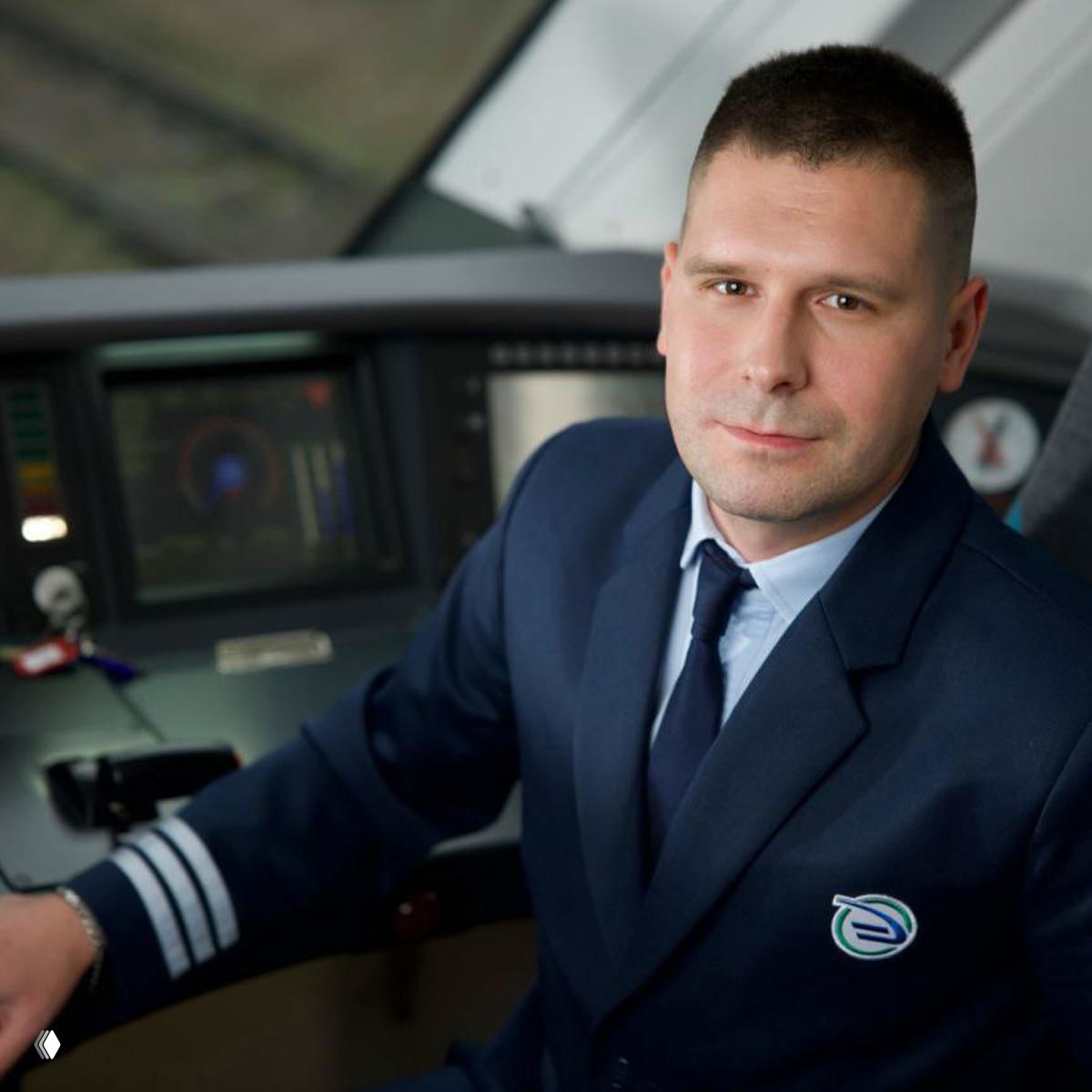 Мужчина в форменной одежде машиниста сидит в кабине электропоезда, видна приборная панель и интерьер кабины управления поезда.