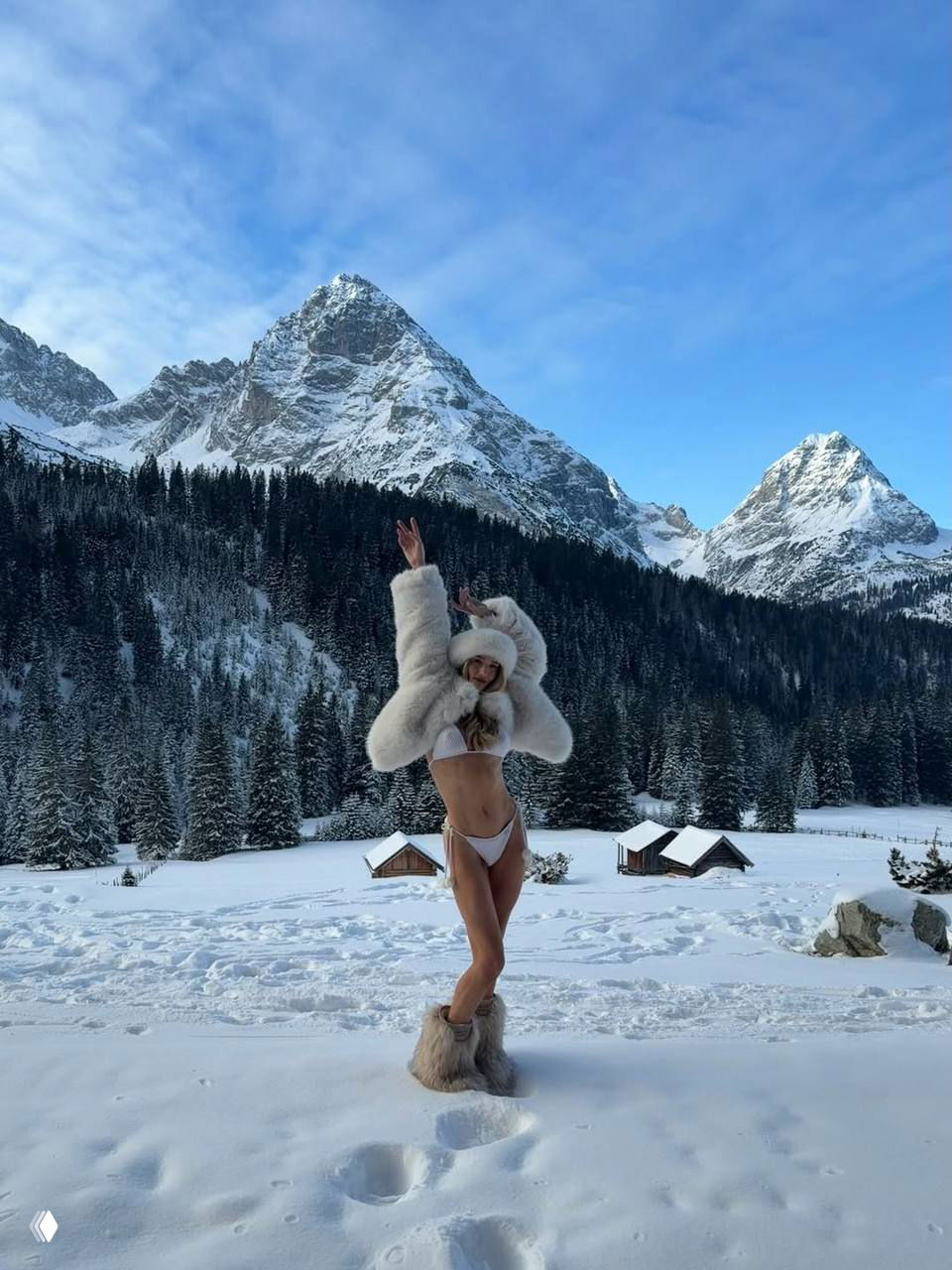 Роузи позирует с поднятыми руками в бикини и меховой шубе на фоне гор и деревянных домиков в снегу — динамичный зимний кадр.