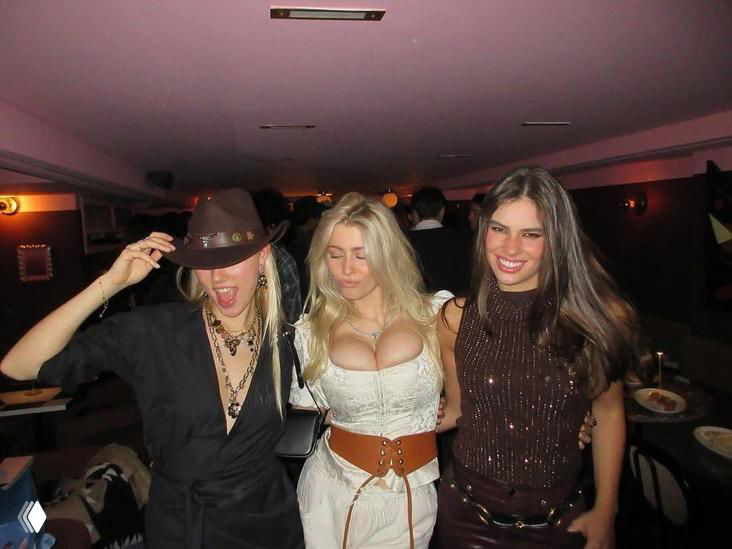 Группа людей на вечеринке: молодая женщина в откровенном платье в ковбойском стиле позирует в помещении с приглушённым светом