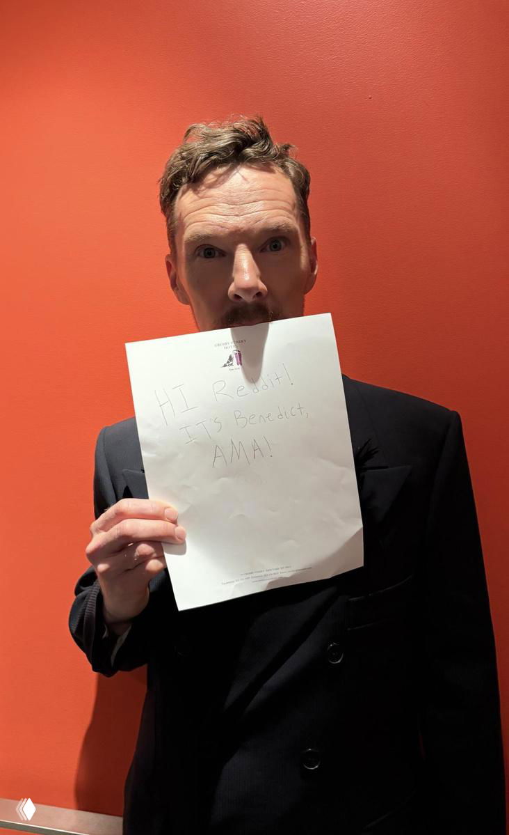 Мужчина в тёмном пиджаке на фоне красной стены держит лист бумаги, частично закрывающий лицо; фото сделано для короткого Q&A с аудиторией.