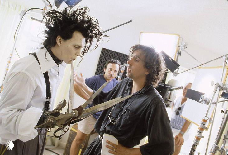 Съёмочный кадр: персонаж с ножницами вместо рук в гриме на площадке, вокруг операторы, свет и реквизит, подготовка сцены.