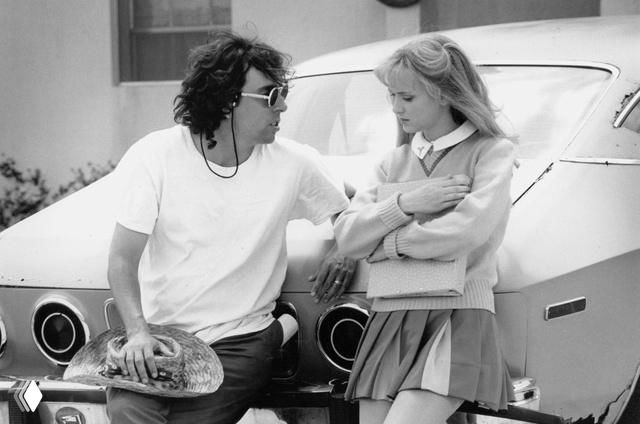 Чёрно‑белый кадр: диалог между членом съёмочной группы и молодой актрисой у автомобиля в гриме и костюме для сцены.