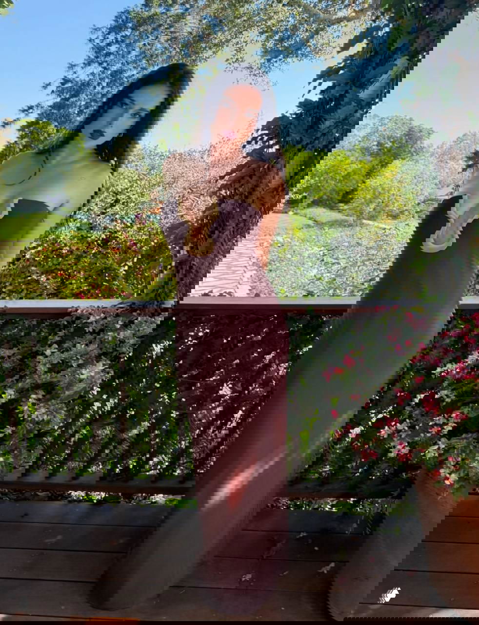 Женщина в длинном коричневом платье стоит на балконе с зелёной садовой растительностью, фото подчёркивает элегантность и позу модели.