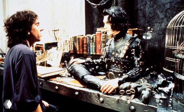 Кадр из сцены в интерьере: персонаж в мрачном костюме сидит за столом в мастерской, вокруг книги и механизмы декора.