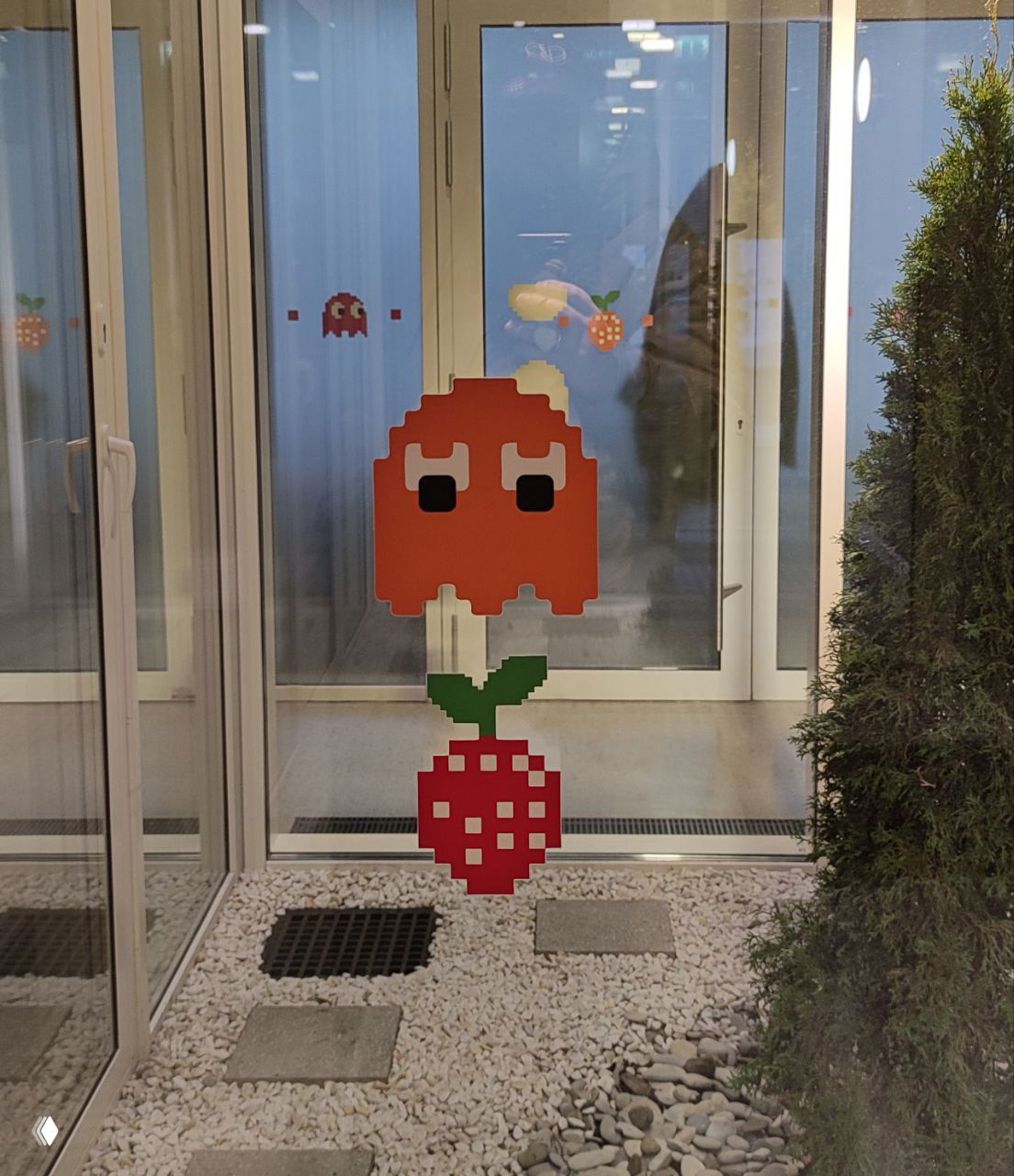 Пиксельная наклейка в виде привидения и клубника на стеклянной перегородке внутреннего дворика кампуса Неймарка, видны отражения коридора.