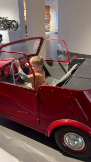 Мальчик сидит в маленьком ярко-красном детском автомобиле в зале музея, вид сбоку, элементы салона и лаковые панели на фоне.