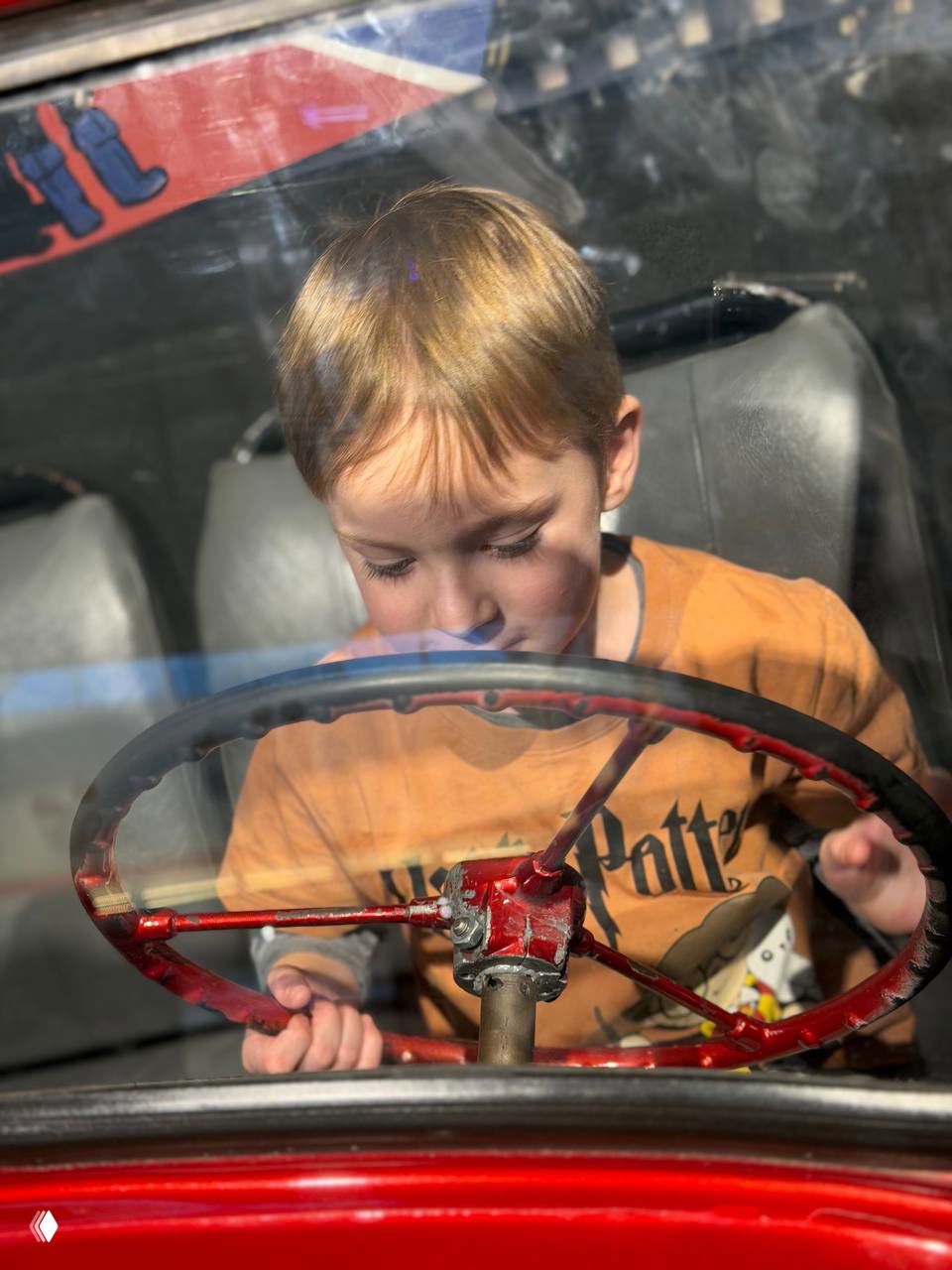 Крупный план ребёнка за рулём старинной машины: лицо ребёнка видно через стекло, красный руль и атмосфера музейной экспозиции вокруг.