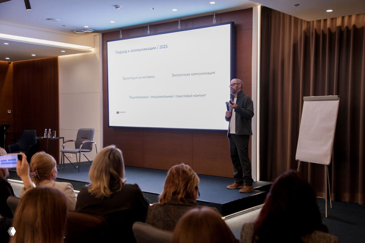 Лектор выступает на сцене перед аудиторией: слайд о коммуникациях и трендах SMM, конференц-зал и флипчарт на сцене.