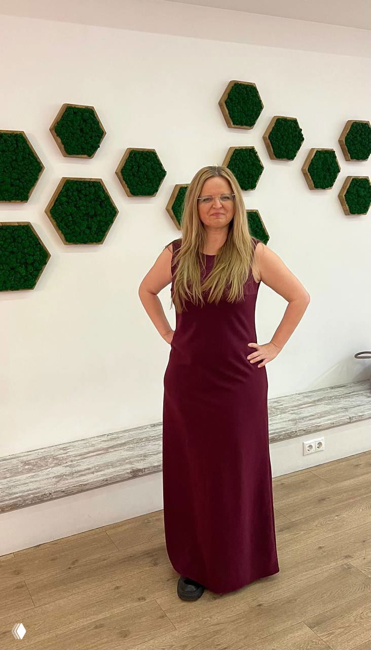 Женщина в длинном бордовом платье стоит перед белой стеной с зелёными шестиугольными декоративными панелями, руки на поясе.